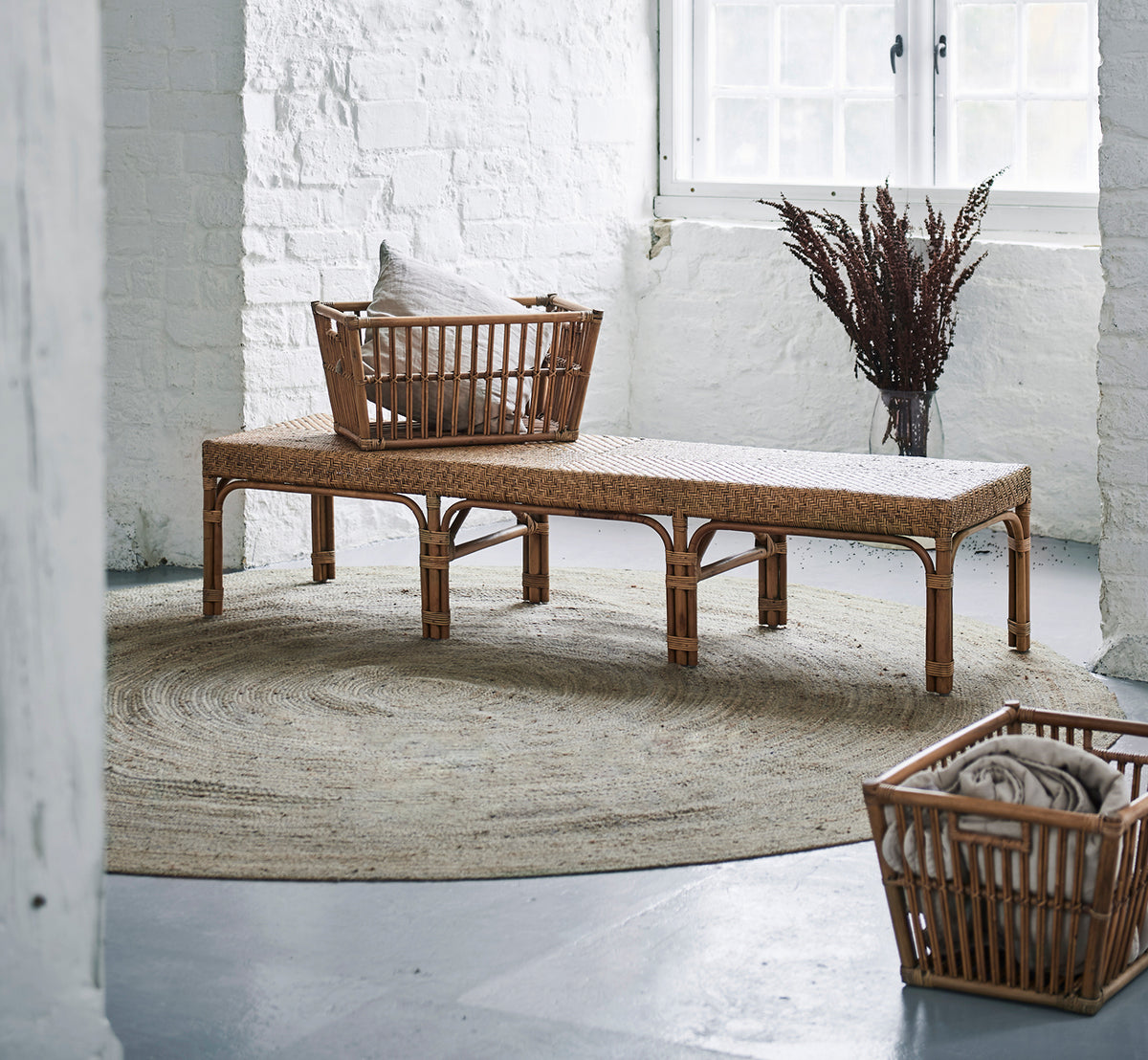 Dies ist ein Lifestyle-Bild von Luis Bank in antique rattan. In einem weißen Backsteinloft mit Korb, Juteteppich und getrockneten Gräsern platziert.