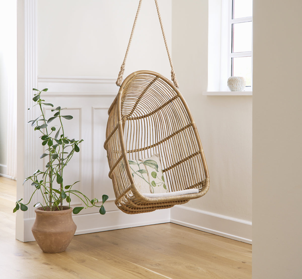 Dies ist ein Lifestyle-Bild von Renoir Hängesessel in antique rattan. In einem hellen vertäfelten Raum neben einer Topfpflanze hängend.