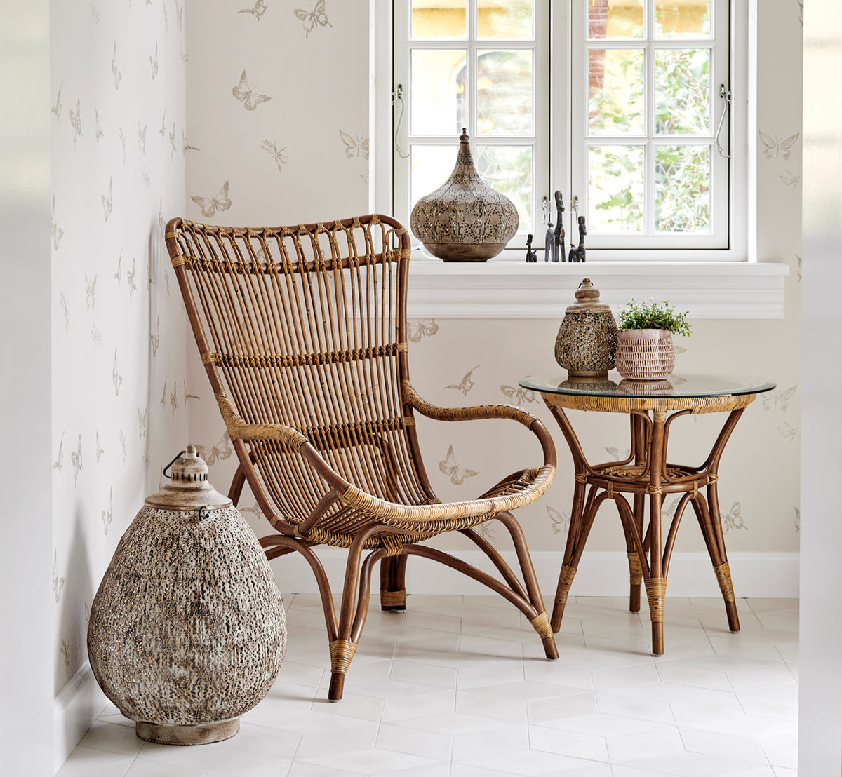 Dies ist ein Lifestyle-Bild von Tony Beistelltisch in antique rattan. In einer mit Schmetterlingsmuster tapezierten Ecke mit Laterne und Deko aufgestellt.