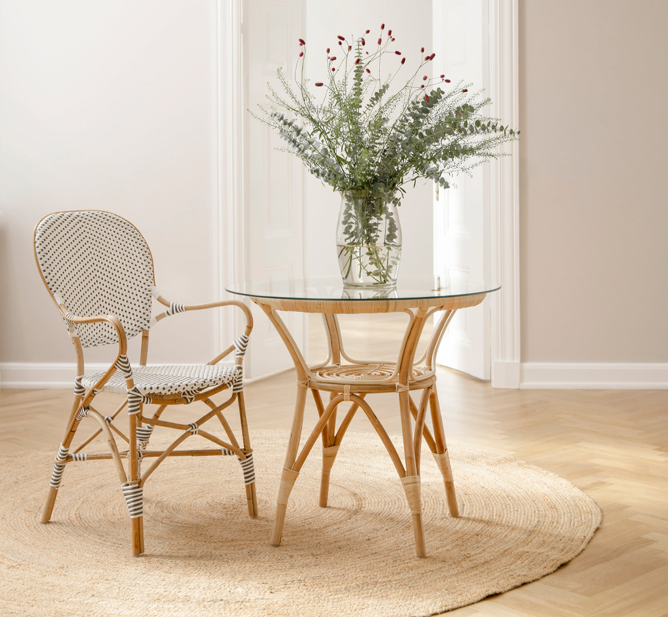Dies ist ein Lifestyle-Bild von Tony Café Table in natural rattan. Mit zwei Stühlen auf einem runden Juteteppich in einem hellen Türrahmen kombiniert.
