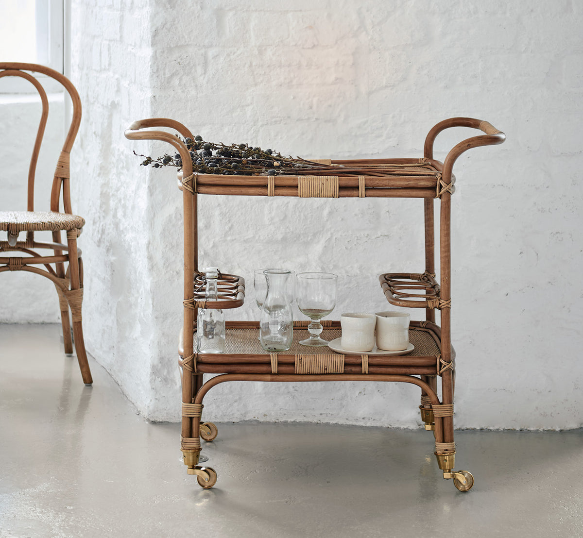 Dies ist ein Lifestyle-Bild von Carlo Trolley in antique Rattan, in einem hell-weißen Interieur mit Wandvertäfelung und einem Rattan-Wandspiegel.