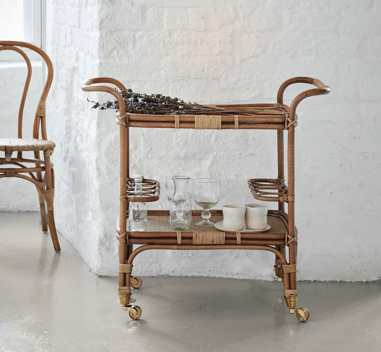 Dies ist ein Lifestyle-Bild von Carlo Trolley in antique Rattan, in einem hell-weißen Interieur mit Wandvertäfelung und einem Rattan-Wandspiegel.