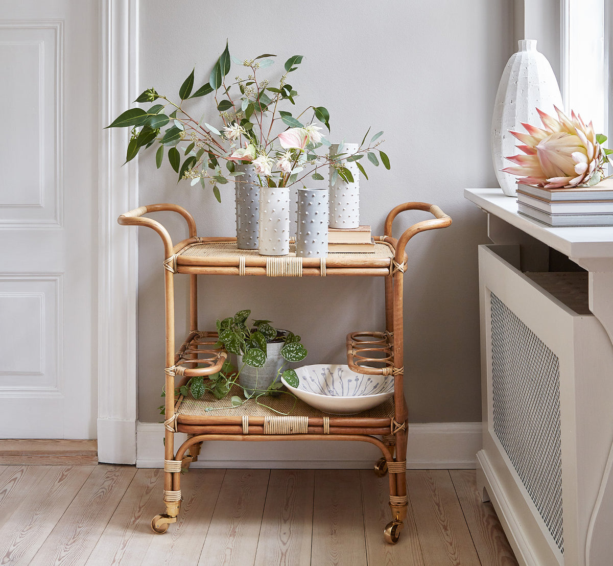 Dies ist ein Lifestyle-Bild von Carlo Trolley in antique Rattan, in einem hell-weißen Interieur mit Wandvertäfelung und einem Rattan-Wandspiegel.