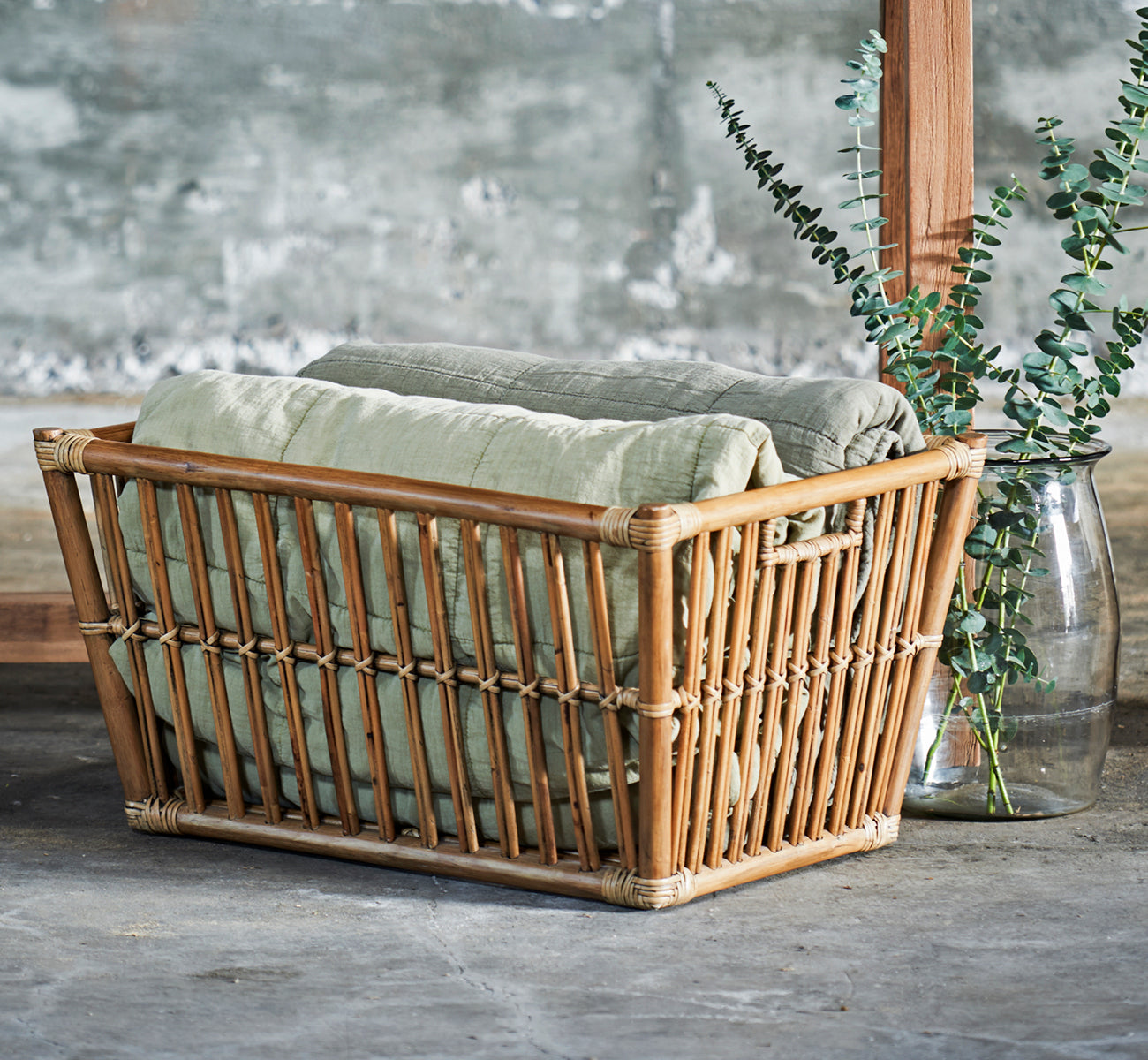 Dies ist ein Lifestyle-Bild von Marche Korb in antique rattan. Gestylt mit Leinendecken und Eukalyptus in einem Raum mit Betonwand.