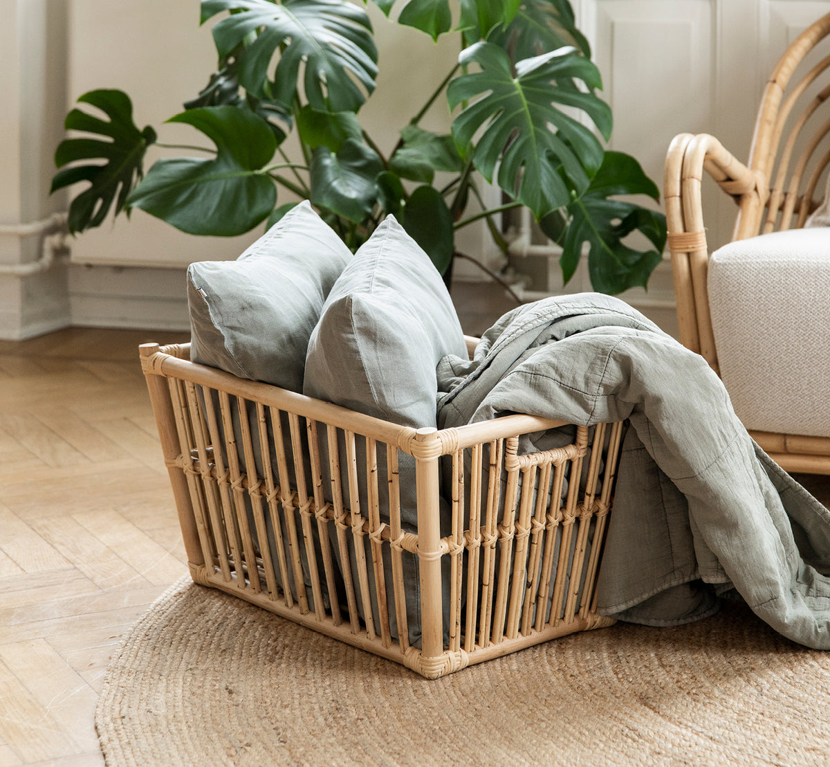 Dies ist ein Lifestyle-Bild von Marche Korb in natural rattan. Gefüllt mit Leinenkissen neben einer Monstera-Pflanze auf Eichenboden.