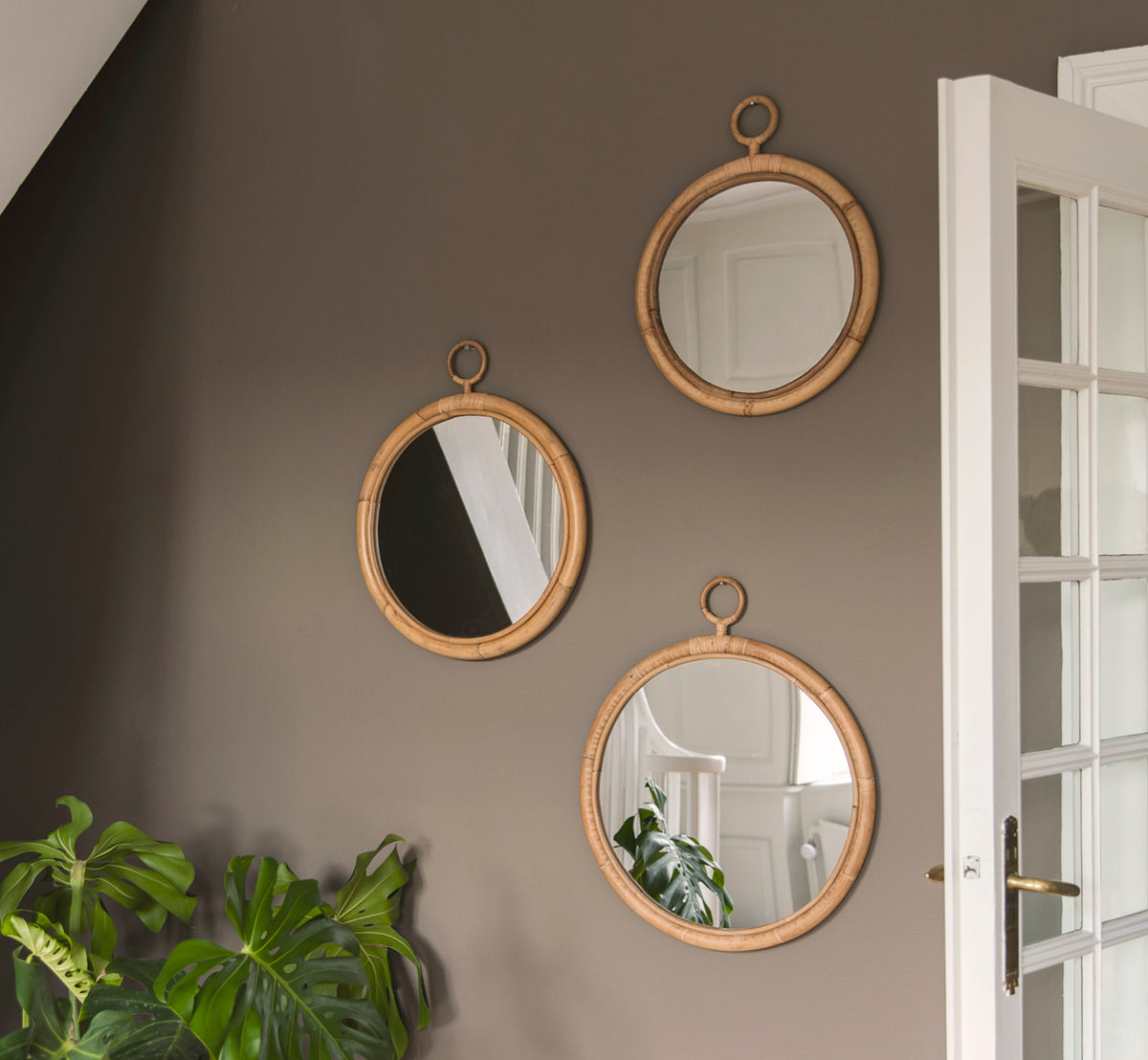 Dies ist ein Lifestyle-Bild von Ella Spiegel Ø45cm in natural skin on rattan, an einer taufarbenen Wand in einem Flur mit einer Monstera-Pflanze und.