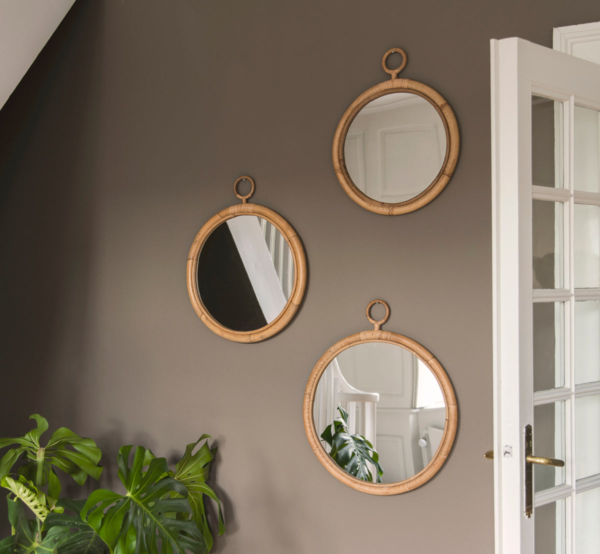 Dies ist ein Lifestyle-Bild von Ella Spiegel Ø50cm in natural skin on rattan, an einer taufarbenen Wand in einem Flur mit einer Monstera-Pflanze und.
