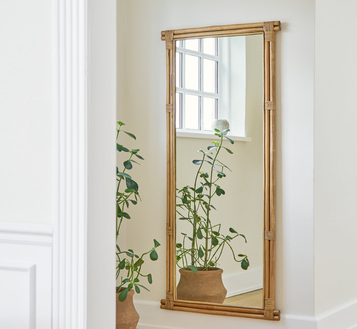 Dies ist ein Lifestyle-Bild von Rasmus Mirror 172 cm in antique rattan. An einer weißen Wand über einer Topfpflanze in einem hellen Korridor montiert.