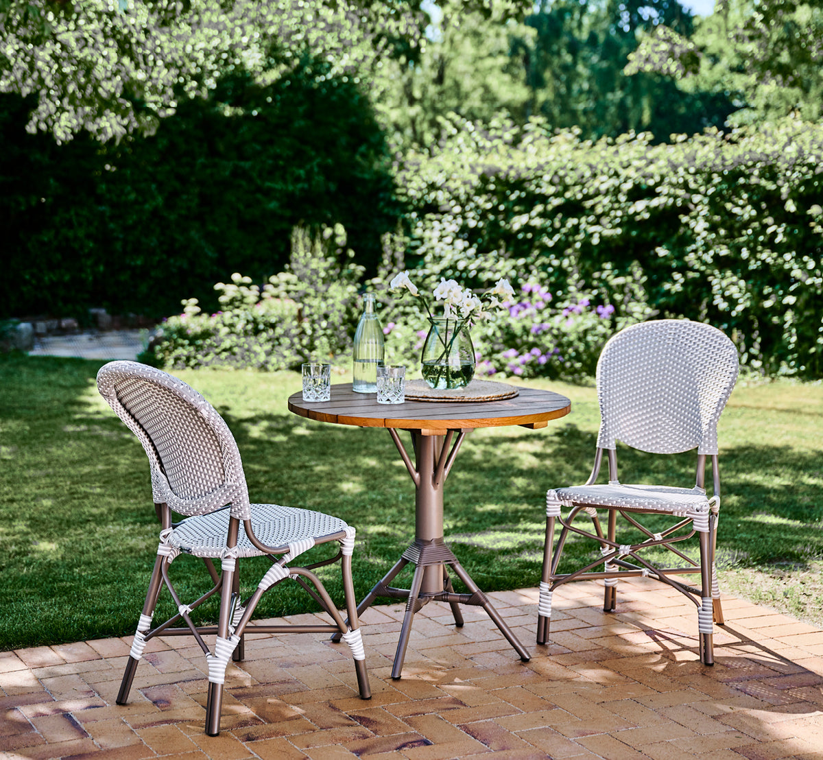 Dies ist ein Lifestyle-Bild von Isabell in taupe aluminium frame Grey w white dot ArtFibre, auf einer üppigen Gartenterrasse mit Steinboden und grün.