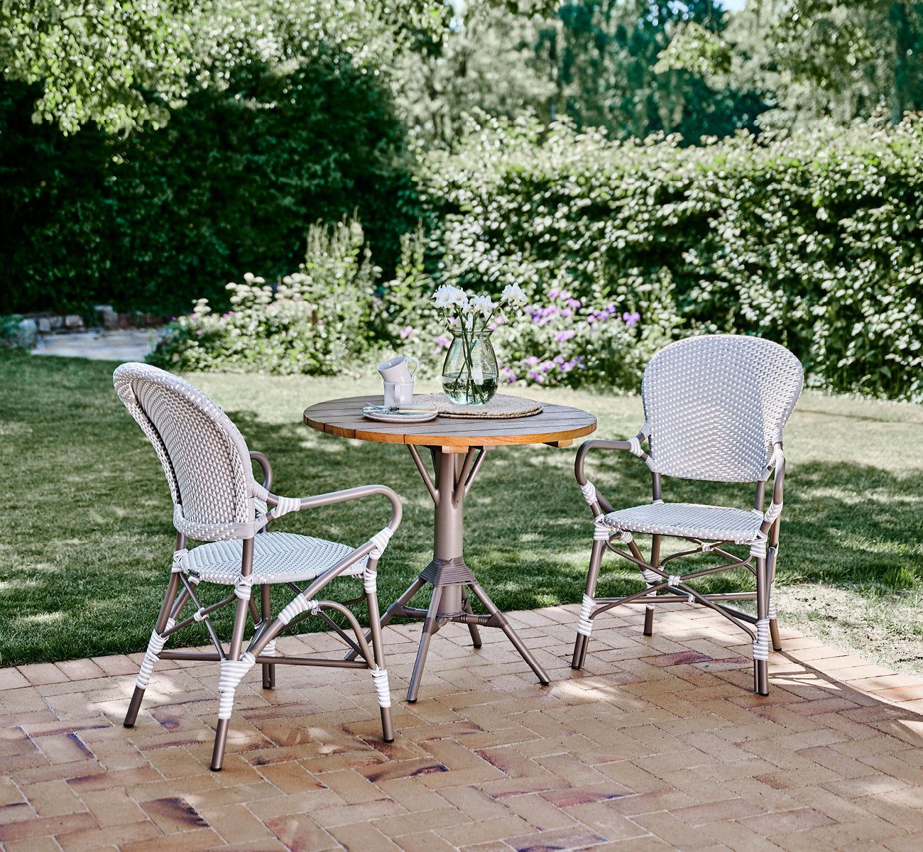 Dies ist ein Lifestyle-Bild von Isabell in taupe aluminium frame Grey w white dot ArtFibre, auf einer üppigen Gartenterrasse mit Steinboden und grün.