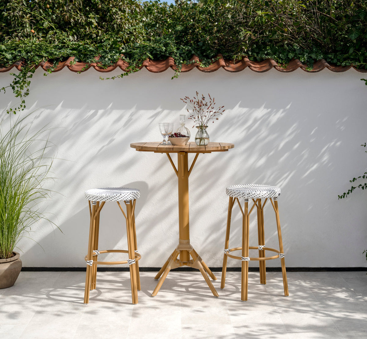 Dies ist ein Lifestyle-Bild von Simone Außen-Barhocker in almond aluminium frame white w cappuccino dot artfibre. An einem Teak-Bartisch auf einer Terrasse.