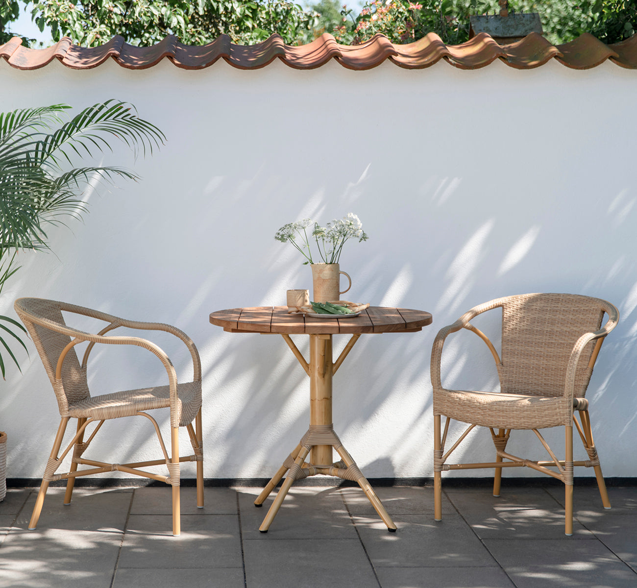 Dies ist ein Lifestyle-Bild von Madeleine Außen-Esszimmerstuhl in almond aluminium frame natural ArtFibre. Innenhof-Bistro mit rundem Teaktisch.