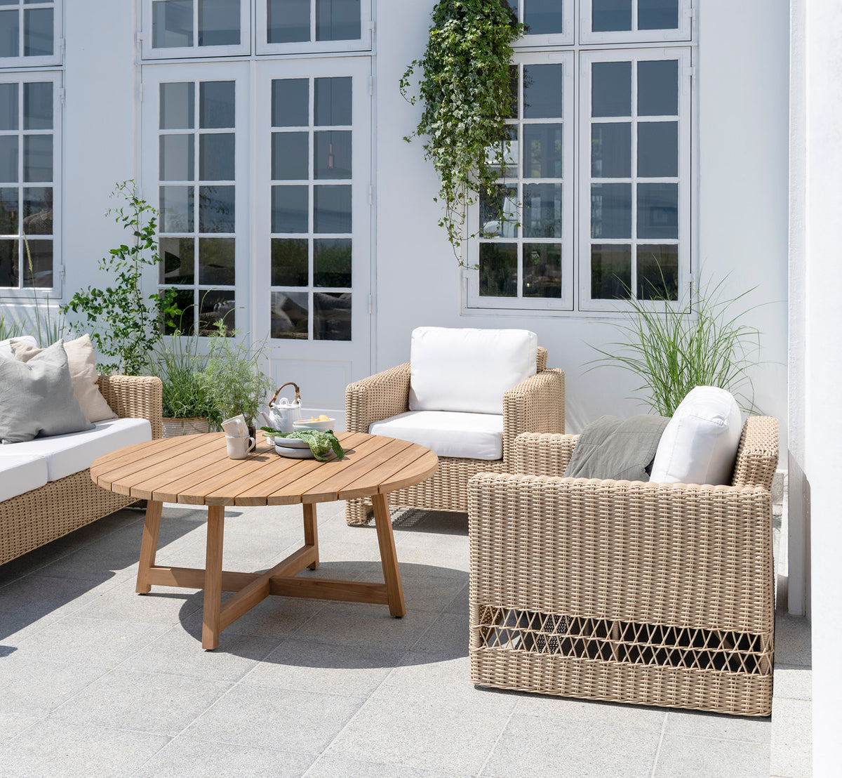 Dies ist ein Lifestyle-Bild von Carrie Lounge-Sessel in natural ArtFibre Aluminium, auf einer sonnendurchfluteten Küstenterrasse mit Meerblick und.