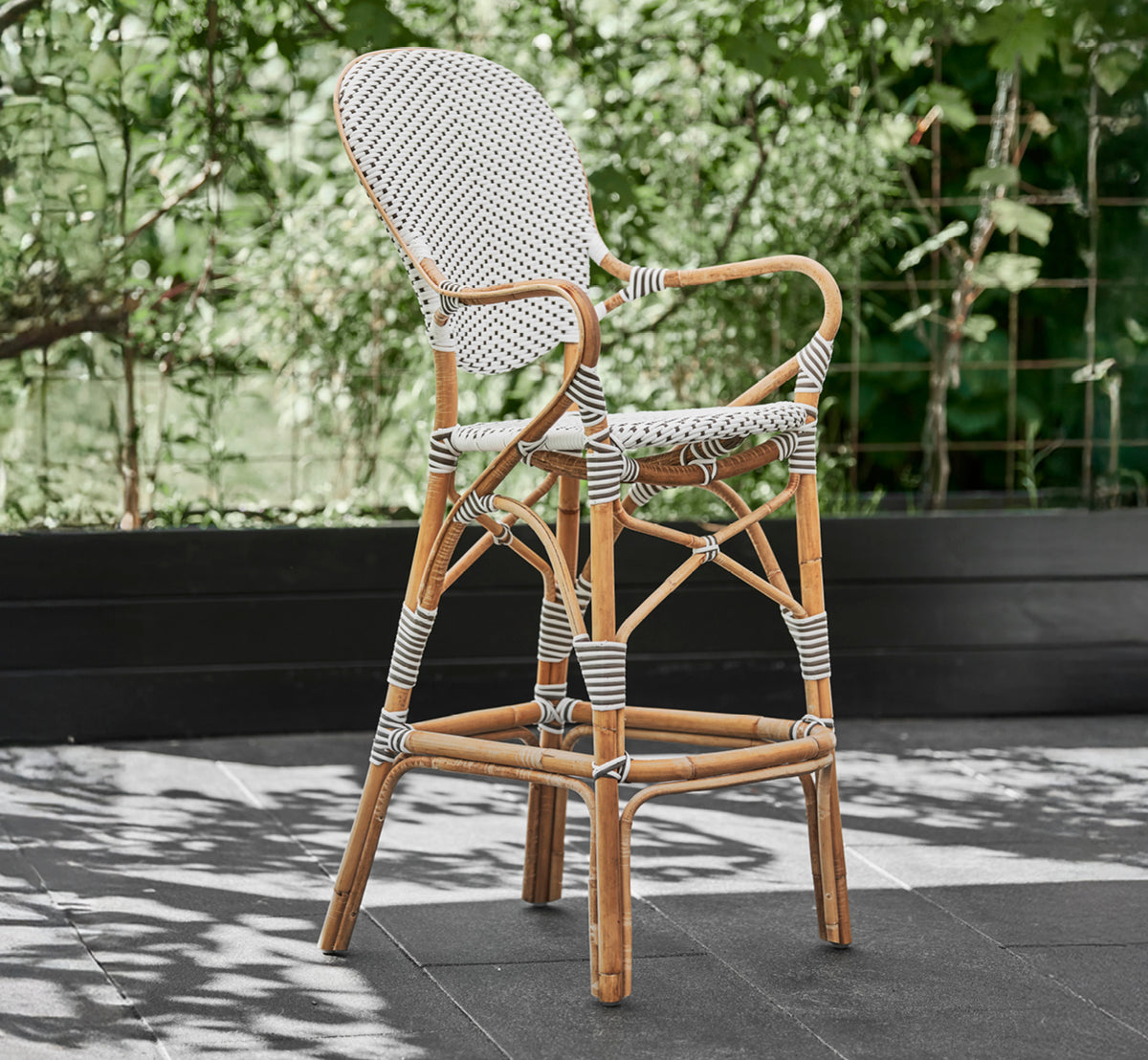 Dies ist ein Lifestyle-Bild von Isabell Barhocker in white w cappuccino dot ArtFibre Skin on Rattan frame, im Freien vor einer üppig grünen.