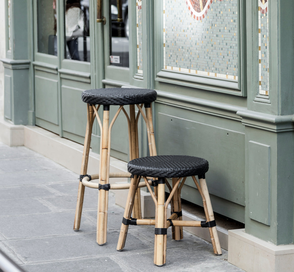 Dies ist ein Lifestyle-Bild von Simone Hocker in black ArtFibre Skin on Rattan frame. Vor einer grünen Pariser Café-Fassade platziert.