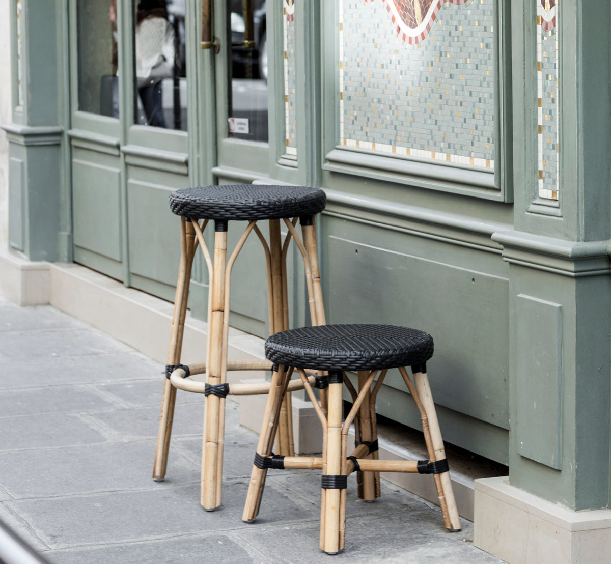 Dies ist ein Lifestyle-Bild von Simone Barstuhl in black ArtFibre Skin on Rattan frame. Vor einer grünen Pariser Café-Fassade platziert.