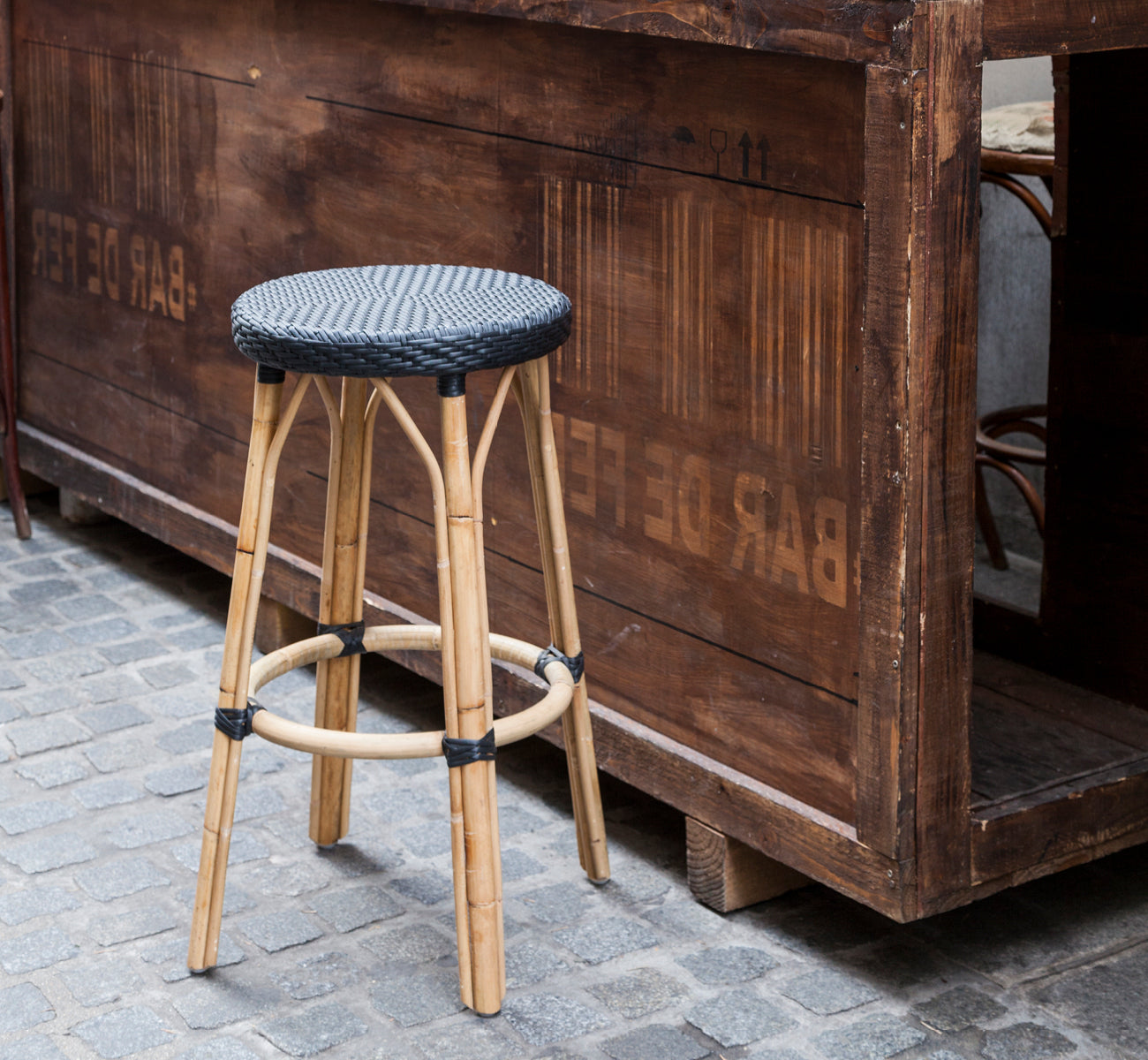 Dies ist ein Lifestyle-Bild von Simone Barhocker in black ArtFibre Skin on Rattan frame. An einer rustikalen Holzbar auf Kopfsteinpflaster.