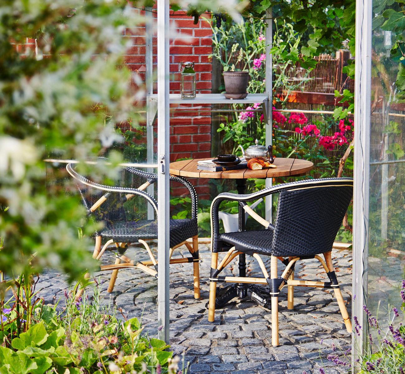 Dies ist ein Lifestyle-Bild von Madeleine Esszimmerstuhl in black ArtFibre skin on rattan frame. Gartengewächshaus mit Kopfsteinpflaster und Pflanzen.