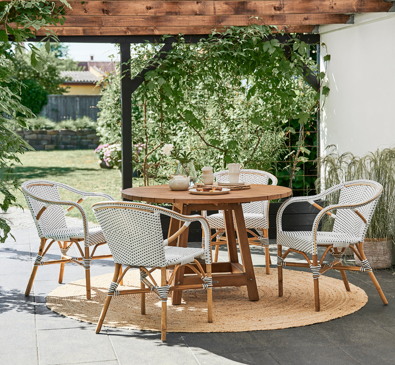 Dies ist ein Lifestyle-Bild von Madeleine Esszimmerstuhl in white w cappuccino dot ArtFibre skin on rattan frame. Gartenpergola mit Juteteppich.