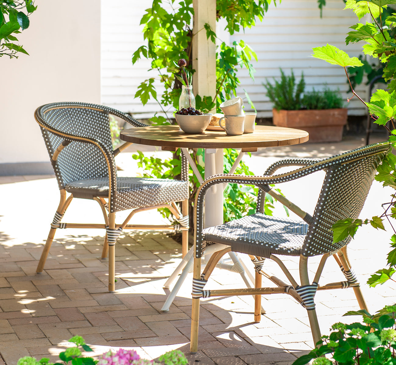 Dies ist ein Lifestyle-Bild von Madeleine Esszimmerstuhl in cappuccino w white dot rattan frame. Schattige Gartenterrasse mit rundem Teaktisch.