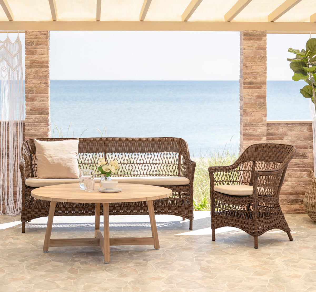 Dies ist ein Lifestyle-Bild von Charlot Außenstuhl in brown artfibre aluminum. Mit Teak-Couchtisch auf einer Terrasse am Meer.