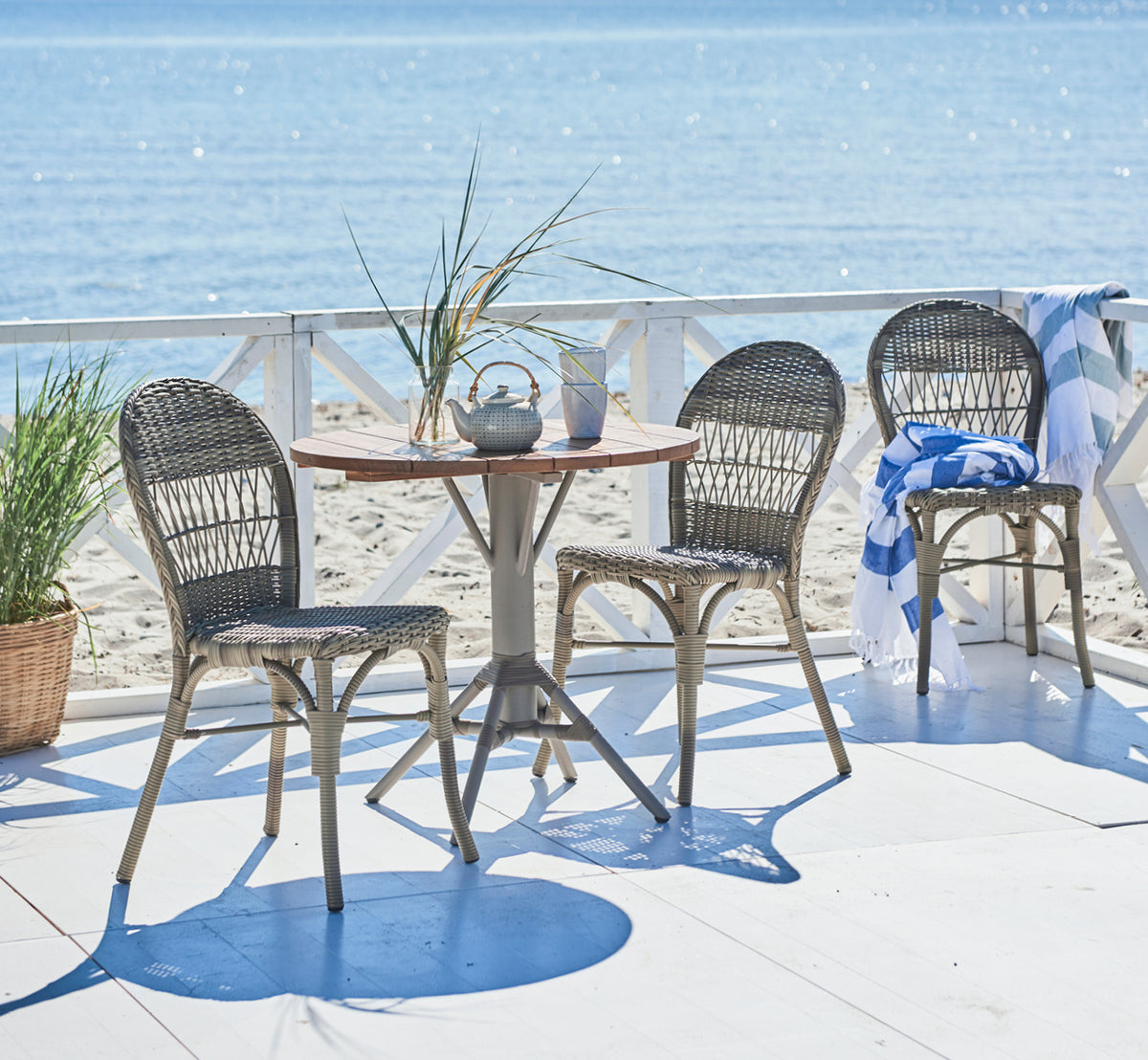 Dies ist ein Lifestyle-Bild von Ofelia Exterior Chair i antique grey artfibre aluminium. Auf einem Deck am Meer mit weißem Geländer und Handtuch.