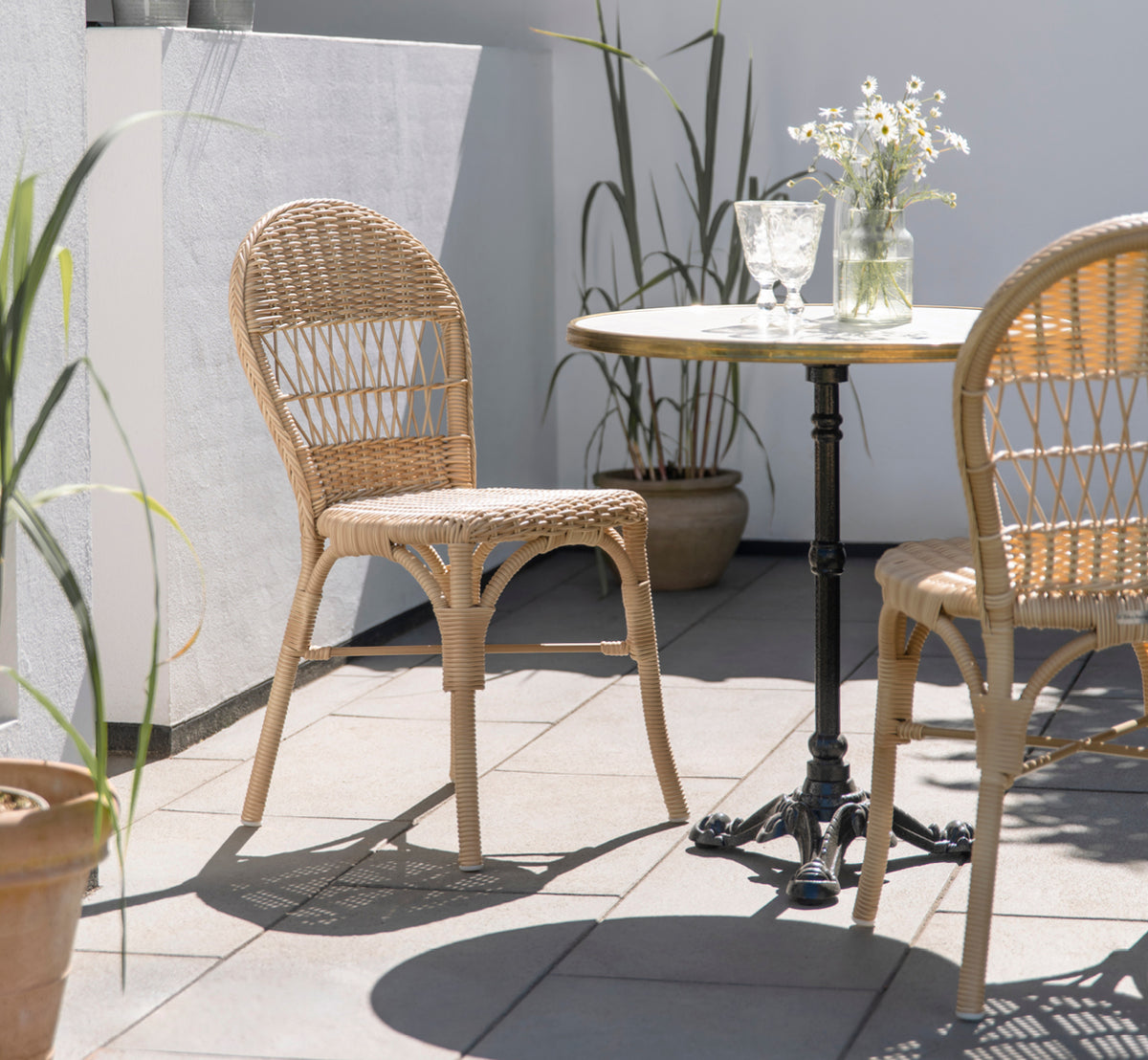 Dies ist ein Lifestyle-Bild von Ofelia Exterior Chair i natural artfibre aluminium. Auf einer sonnigen Terrasse mit Gänseblümchen und Bistrotisch.