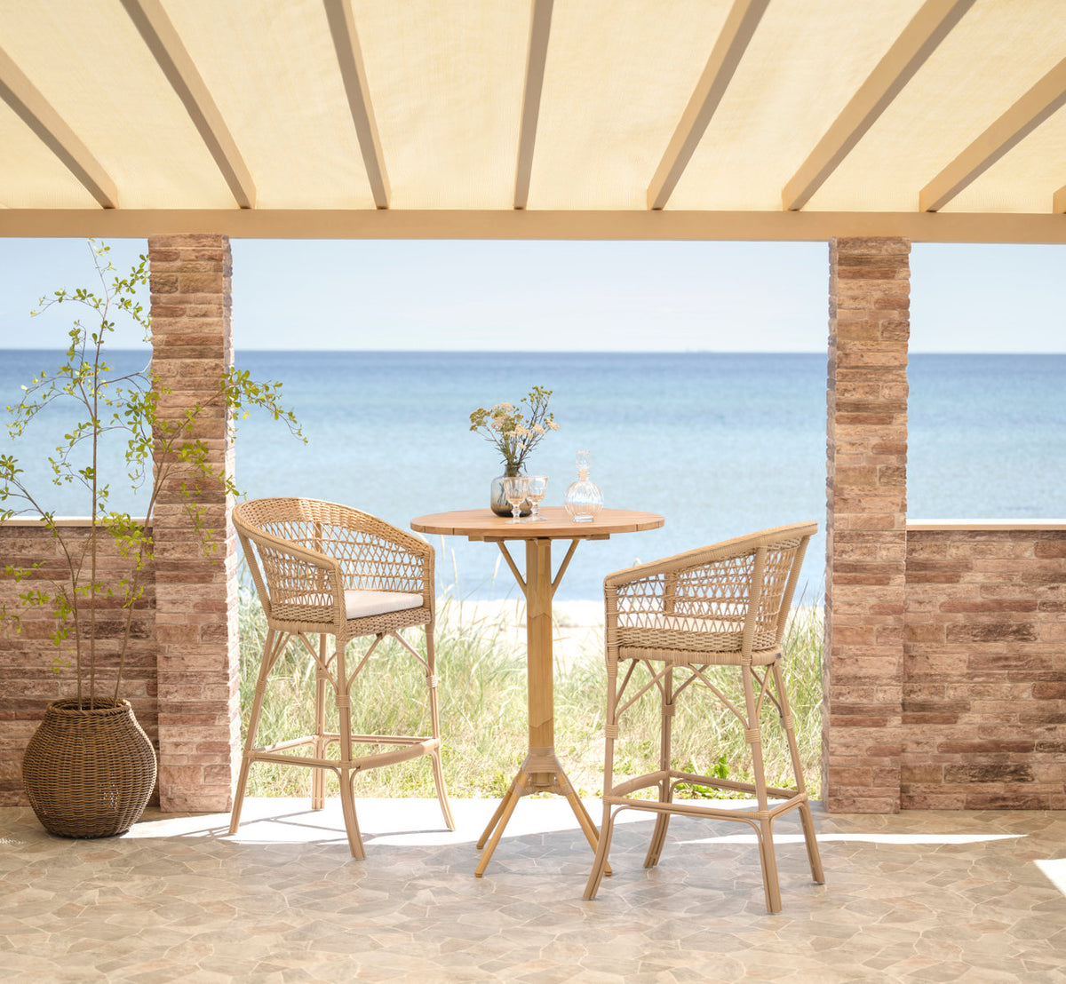 Dies ist ein Lifestyle-Bild von Emma Außen-Barhocker in natural artfibre aluminium. Bar-Ensemble auf einer Terrasse am Meer.