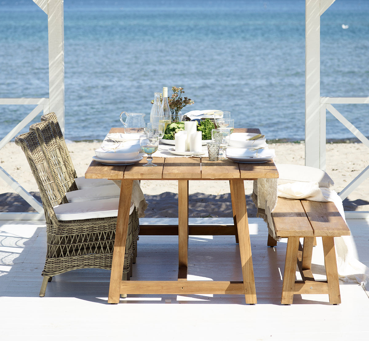 Dies ist ein Lifestyle-Bild von Marie Exterior Side Chair i antique grey artfibre aluminium. An einem Teaktisch in einem Pavillon am Meer.