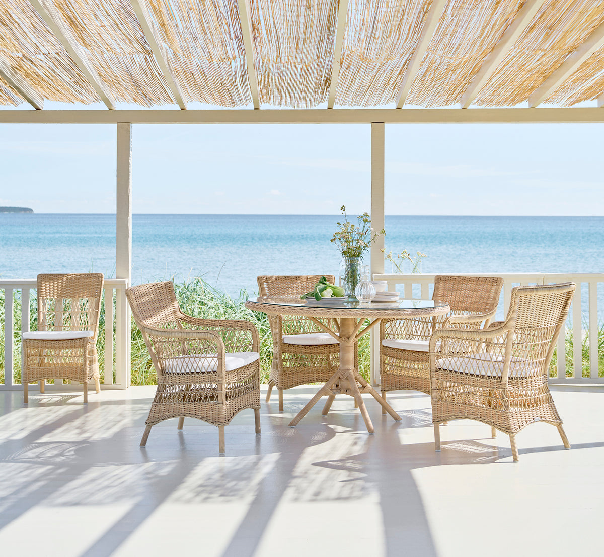 Dies ist ein Lifestyle-Bild von Marie Exterior Side Chair i natural artfibre aluminium. Um einen Tisch auf einer schattigen Veranda am Meer.