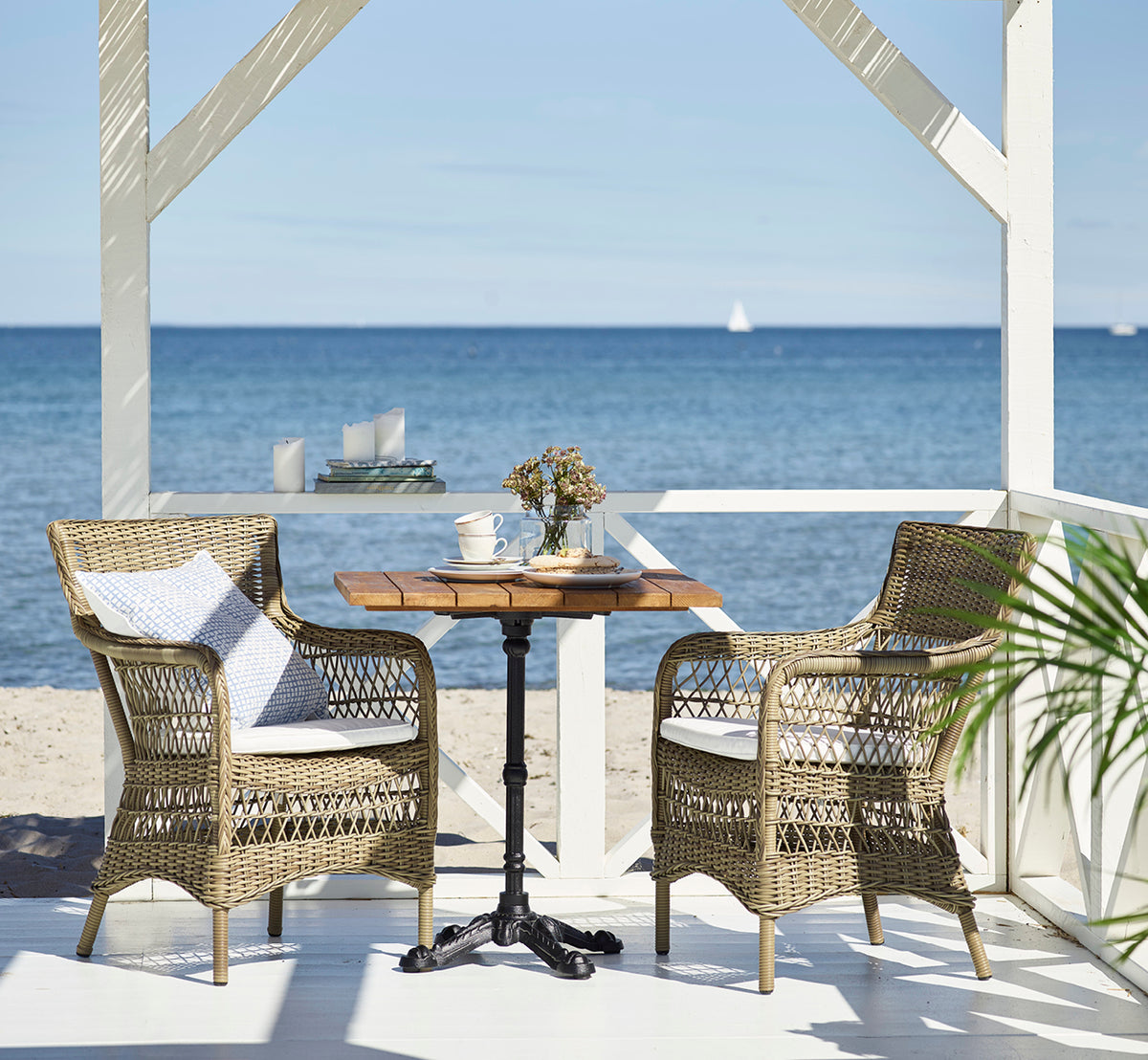 Dies ist ein Lifestyle-Bild von Marie Exterior Armchair i antique grey artfibre aluminium. An einem Teaktisch in einem Strandpavillon.
