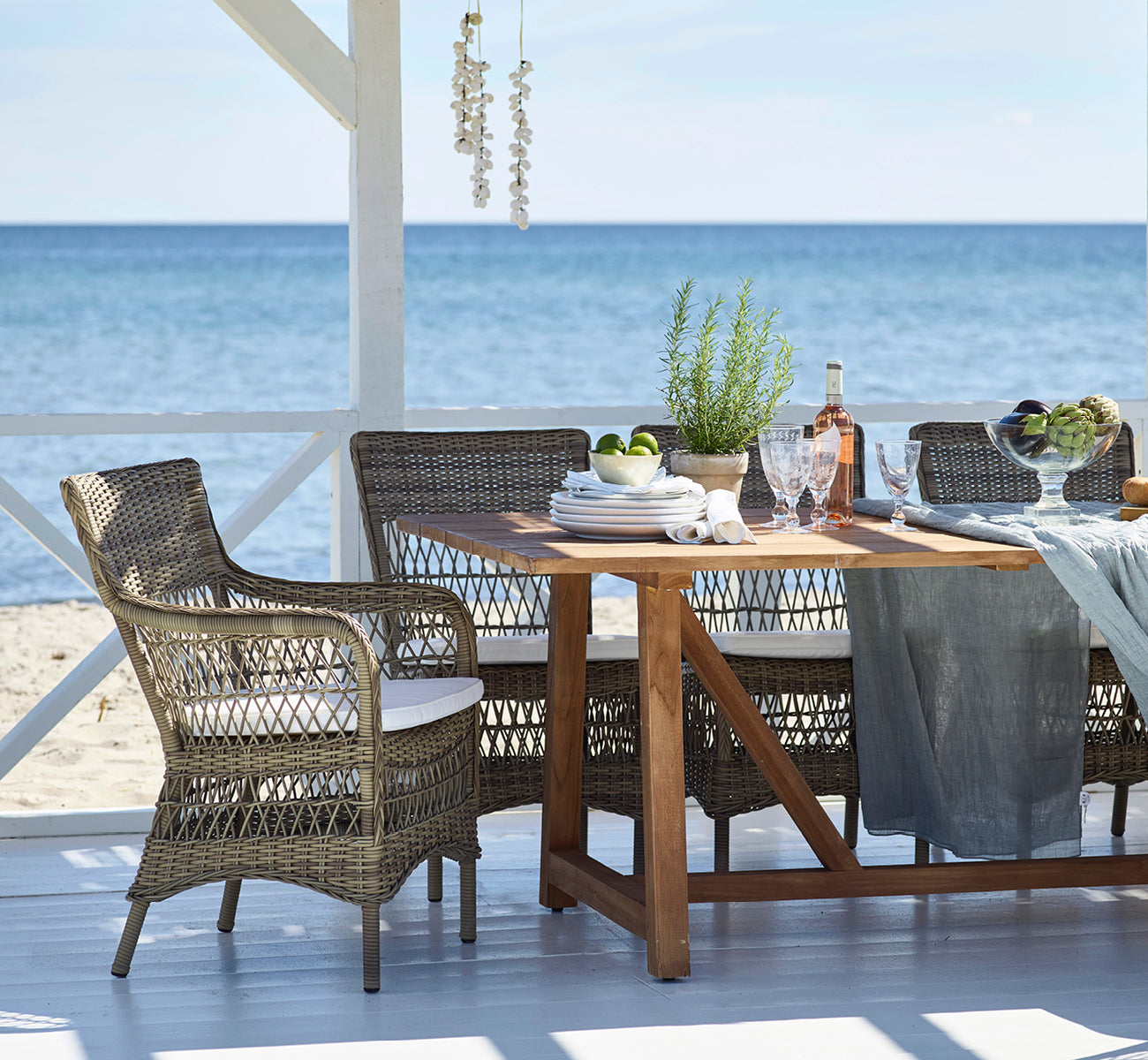 Dies ist ein Lifestyle-Bild von Marie Exterior Armchair i antique grey artfibre aluminium. Um einen Teaktisch in einem Strandpavillon.