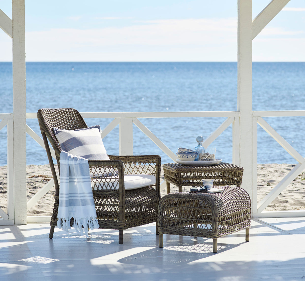 Dies ist ein Lifestyle-Bild von Dawn Lounge-Sessel in antique Grey ArtFibre Aluminium, auf einer Küstenterrasse mit Meerblick, Strandgräsern und.