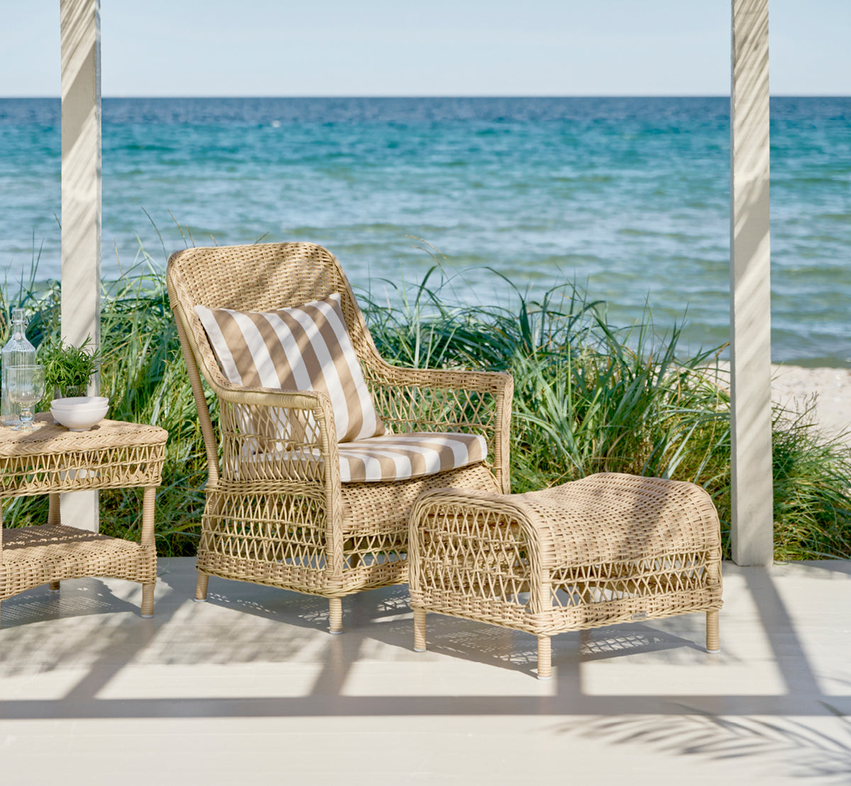 Dies ist ein Lifestyle-Bild von Dawn Lounge-Sessel in natural ArtFibre Aluminium, auf einer Küstenterrasse mit Meerblick, Strandgräsern und weißen.