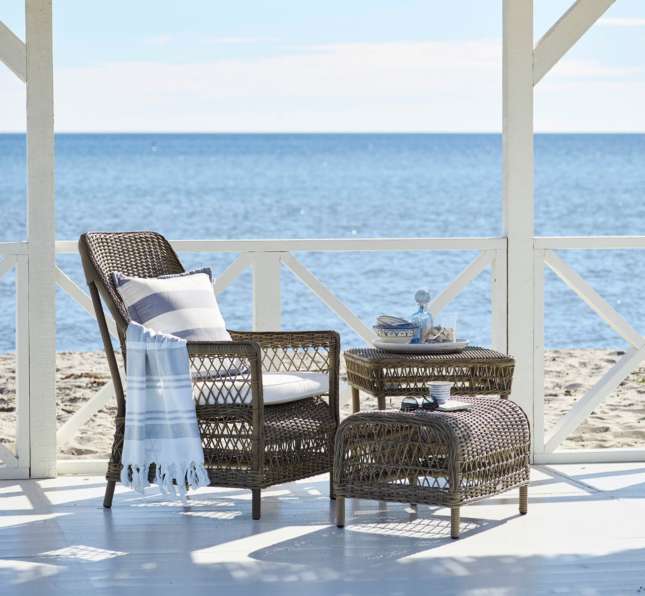 Dies ist ein Lifestyle-Bild von Dawn Fußhocker in antique Grey ArtFibre Aluminium, auf einer Küstenterrasse mit Meerblick, Strandgräsern und weißen.