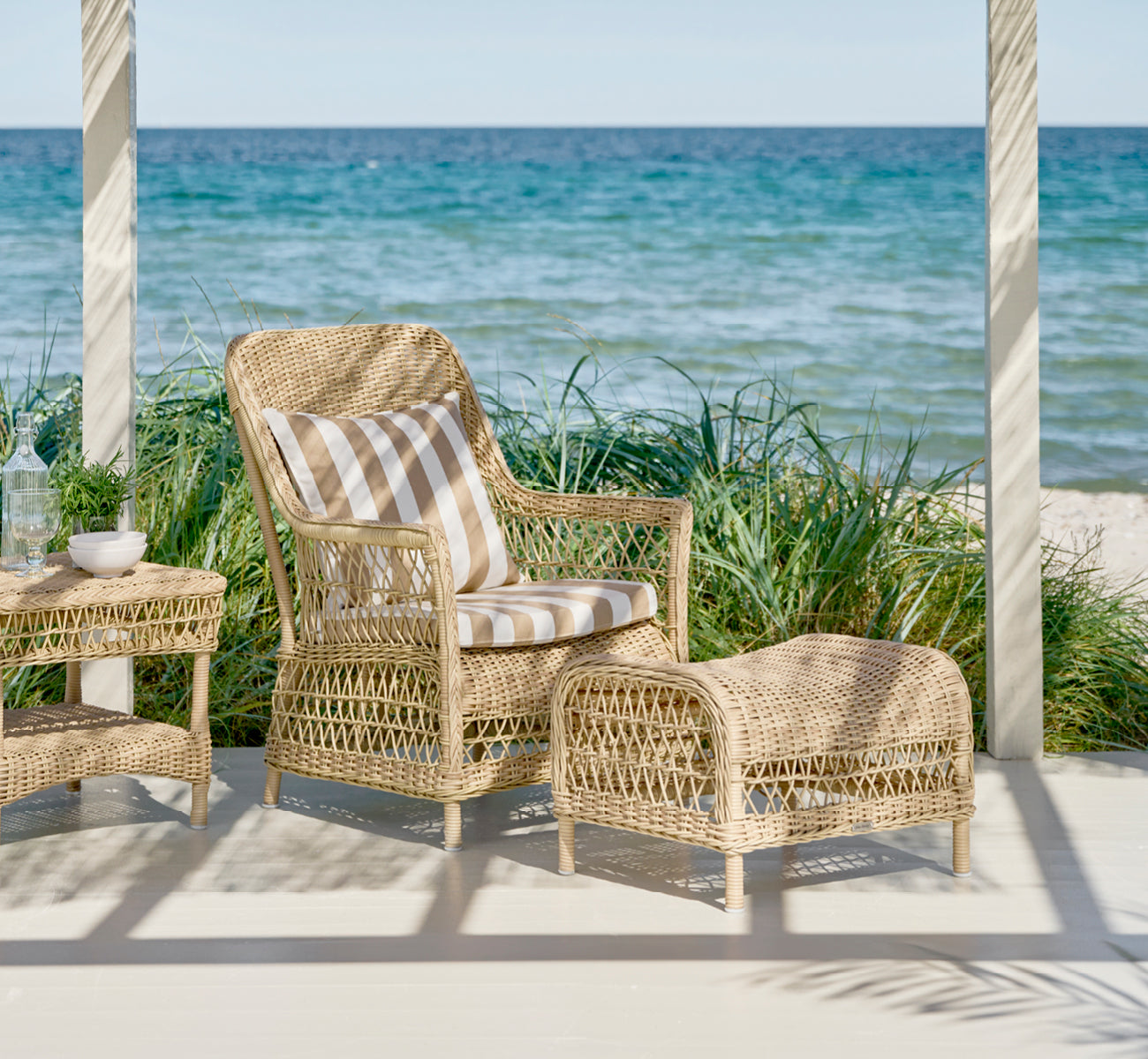Dies ist ein Lifestyle-Bild von Dawn Fußhocker in natural ArtFibre Aluminium, auf einer Küstenterrasse mit Meerblick, Strandgräsern und weißen Säulen.