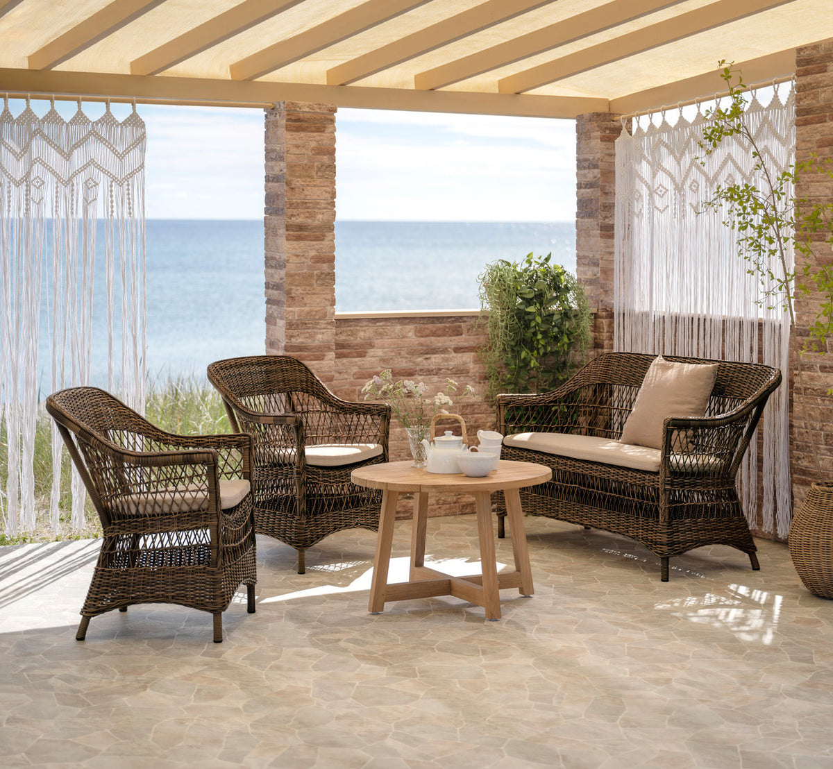 Dies ist ein Lifestyle-Bild von Charlot 2-Sitzer-Außensofa in brown artfibre aluminum. Mit Teak-Couchtisch und Makramee-Vorhängen am Meer.