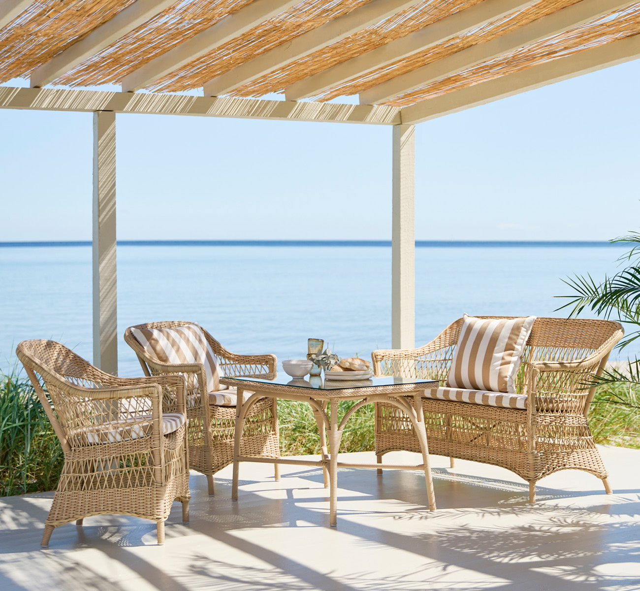 Dies ist ein Lifestyle-Bild von Charlot 2-Sitzer-Sofa in natural ArtFibre Aluminium, auf einer Küstenterrasse mit Schilfdach, Meerblick und.