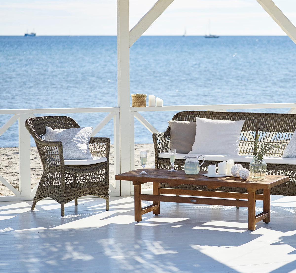 Dies ist ein Lifestyle-Bild von Charlot 3-Sitzer-Sofa in antique Grey ArtFibre Aluminium, auf einer Küstenterrasse mit Schilfdach und Meerblick.