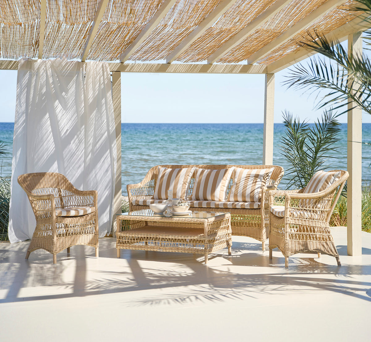 Dies ist ein Lifestyle-Bild von Charlot 3-Sitzer-Sofa in natural ArtFibre Aluminium, auf einer Küstenterrasse mit Schilfdach und Meerblick.