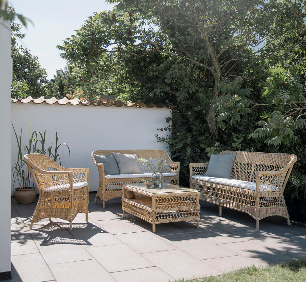 Dies ist ein Lifestyle-Bild von Charlot 3-Sitzer-Sofa in natural ArtFibre Aluminium, auf einer Küstenterrasse mit Schilfdach und Meerblick.