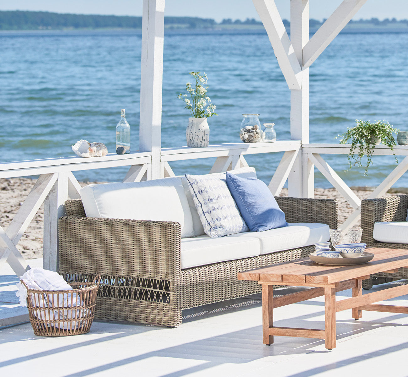 Dies ist ein Lifestyle-Bild von Carrie Sofa in antique Grey ArtFibre Aluminium, auf einer Küstenterrasse mit weißem Holzdeck, Meerblick und Pergola.