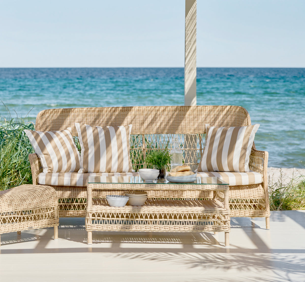 Dies ist ein Lifestyle-Bild von Dawn Sofa in natural ArtFibre Aluminium, auf einer Küstenterrasse mit weißem Holzdeck und Meerblick.