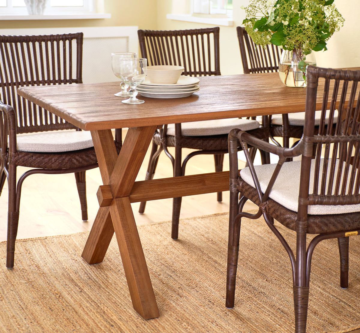 Dies ist ein Lifestyle-Bild von Hugo Teak-Esstisch 180x90 cm in teak indoor. Mit Rattan-Esszimmerstühlen auf einem Juteteppich.