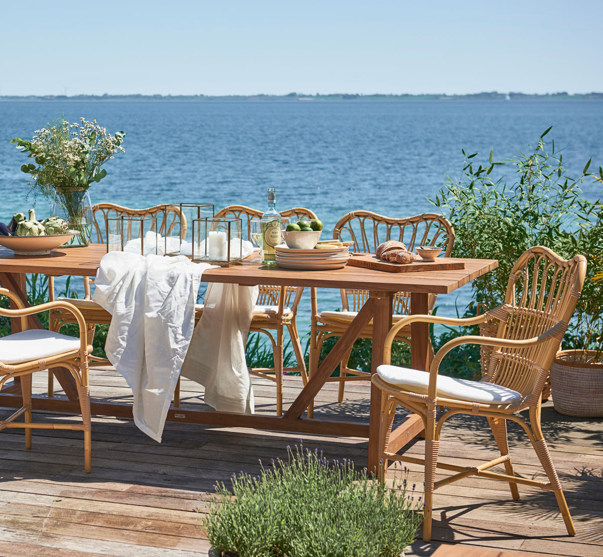 Dies ist ein Lifestyle-Bild von George Außentisch 240 in outdoor Teak, auf einer Gartenterrasse im Freien mit weißer Hausfassade und Bepflanzung.
