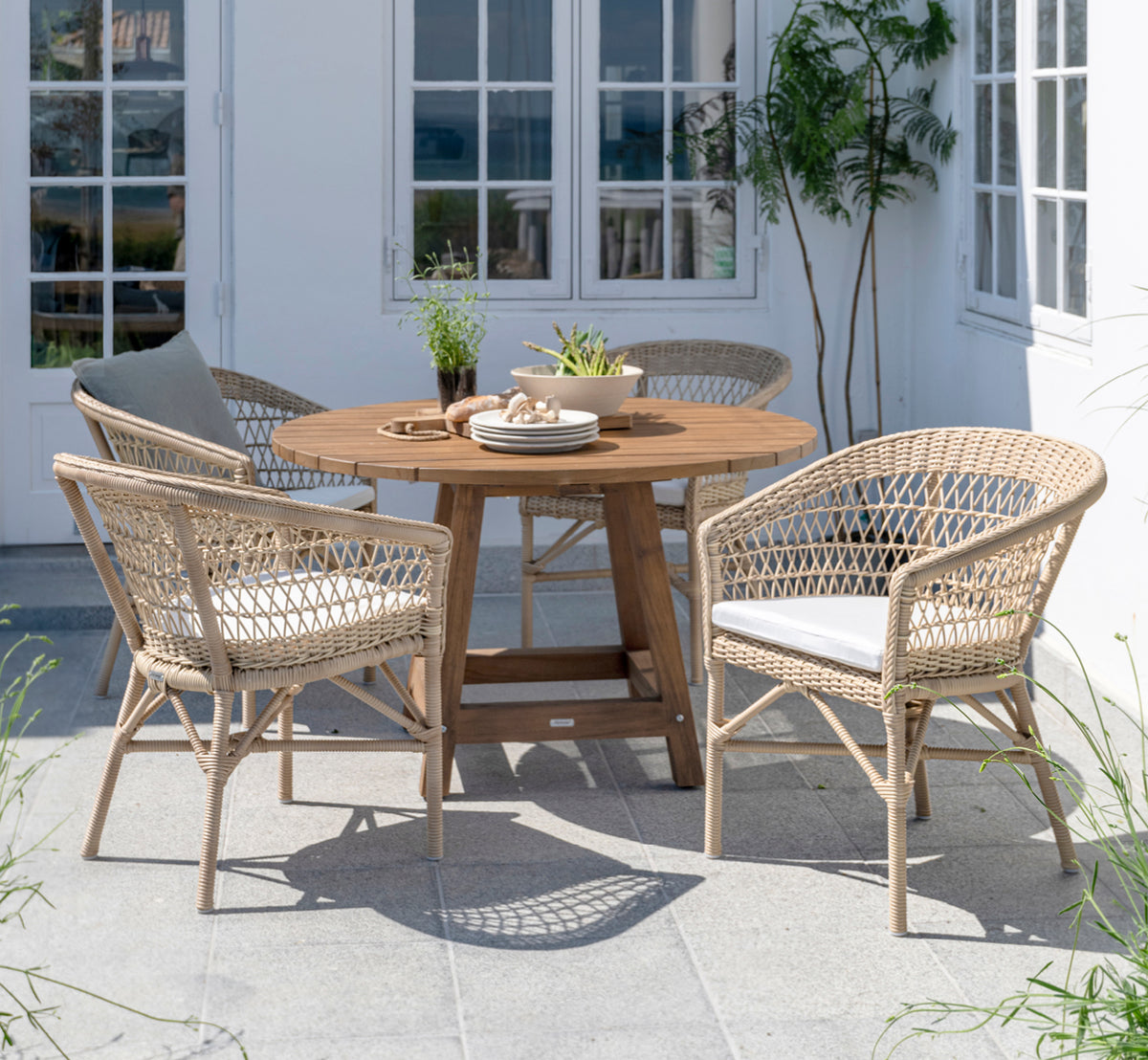 Dies ist ein Lifestyle-Bild von George Außentisch Ø120 in outdoor Teak, auf einer Gartenterrasse im Freien mit weißer Hausfassade und Bepflanzung.