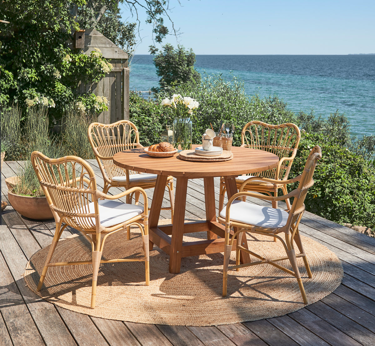 Dies ist ein Lifestyle-Bild von George Außentisch Ø120 in outdoor Teak, auf einer Gartenterrasse im Freien mit weißer Hausfassade und Bepflanzung.