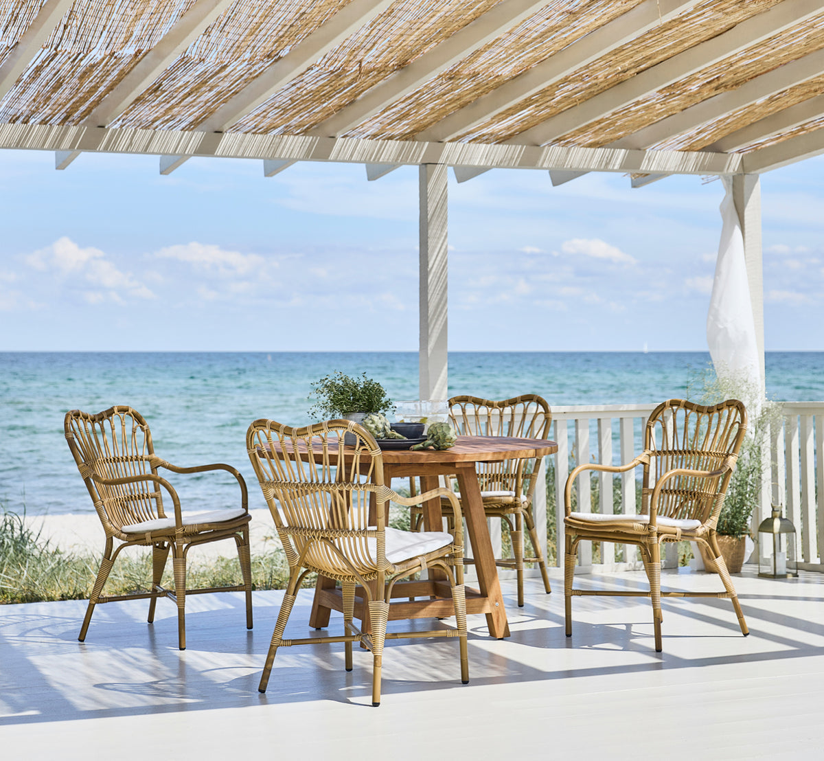 Dies ist ein Lifestyle-Bild von George Außentisch Ø120 in outdoor Teak, auf einer Gartenterrasse im Freien mit weißer Hausfassade und Bepflanzung.