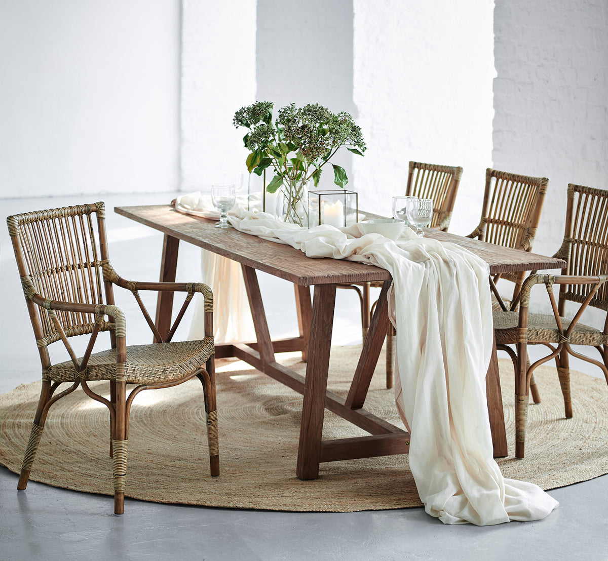Dies ist ein Lifestyle-Bild von Lucas Teak Dining Table 240X100 cm in teak indoor. Mit Leinen und Grün drapiert, umgeben von Rattanstühlen.