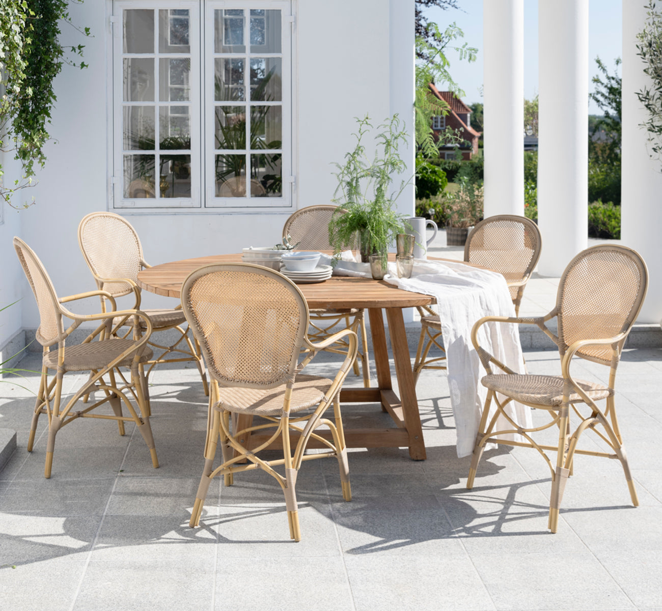 Dies ist ein Lifestyle-Bild von George Außentisch Ø160 in outdoor Teak, auf einer Gartenterrasse im Freien mit weißer Hausfassade und Bepflanzung.